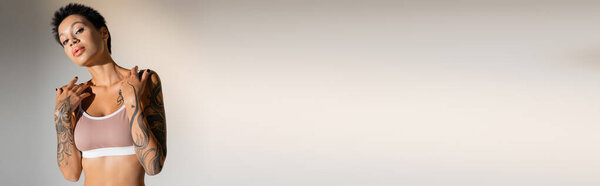 молодая татуированная женщина в лифчике смотрит в камеру на сером фоне, баннер