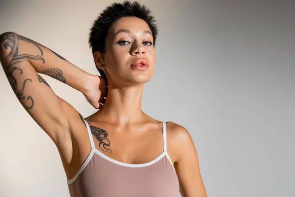young brunette woman with makeup and tattooed body posing with hand behind neck on grey background