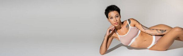 сексуальная брюнетка с татуированным телом лежащим в нижнем белье и смотрящим на камеру на сером фоне, баннер