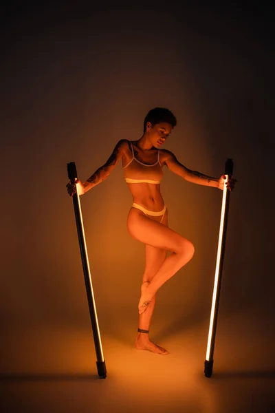 stock image full length of slim tattooed woman in underwear posing with bright fluorescent lamps on dark background