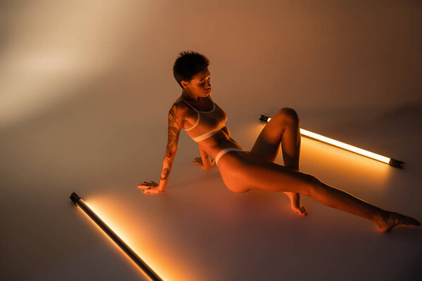 full length of brunette woman with tattooed body sitting in lingerie near fluorescent lamps on beige background