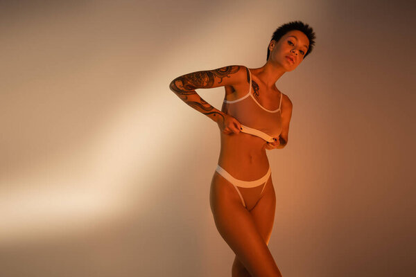young tattooed woman with slender body adjusting bra and looking at camera on beige background