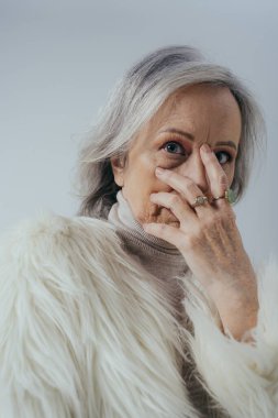 portrait of senior woman with rings on fingers looking at camera while covering face isolated on grey 