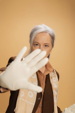 senior woman in white glove looking at camera while showing stop sign isolated on beige 