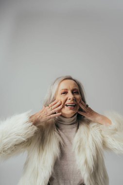 portrait of cheerful senior woman with rings on fingers looking at camera while touching face on grey clipart