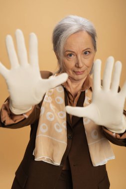 Takım elbiseli ve fularlı yaşlı bir kadın bej rengi eldivenler içinde ellerini gösterirken kameraya bakıyor. 
