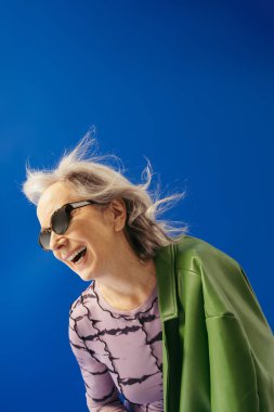 happy senior woman in trendy sunglasses and green leather jacket laughing against wind isolated on blue  clipart