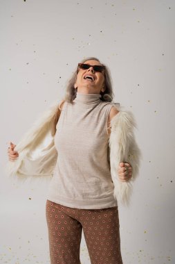 happy elderly woman in white faux fur jacket and sunglasses looking at falling confetti on grey background 
