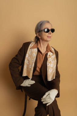 senior woman in brown suit and sunglasses sitting on wooden chair on beige 