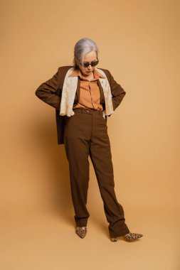 full length of stylish elderly woman in sunglasses and suit posing with hands on hips on beige 