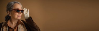 senior woman in brown suit and white gloves adjusting trendy sunglasses on beige, banner 