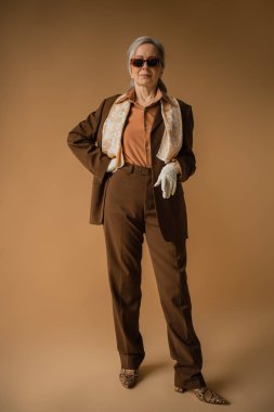 full length of senior woman in brown suit and white gloves posing with hand on hip on beige 