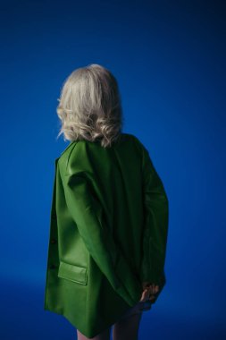 back view of grey haired woman in green leather jacket standing with hands behind back isolated on blue clipart