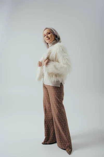 stock image full length of senior woman in faux fur jacket posing on grey background  