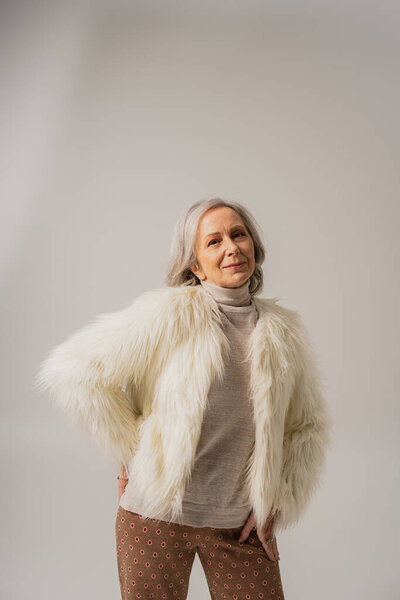pleased elderly woman in white faux fur jacket posing with hand on hip on grey 