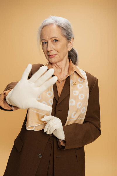 elderly woman in white gloves looking at camera while showing stop sign isolated on beige 
