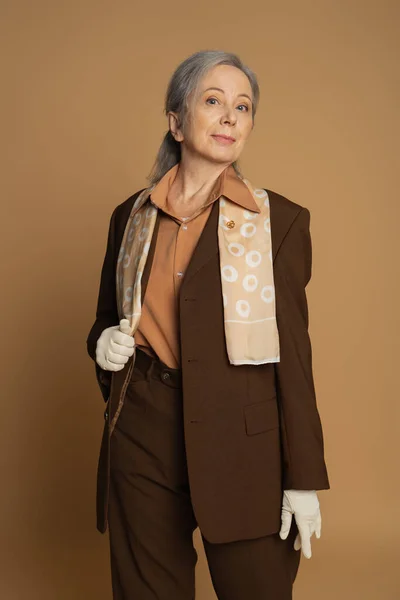 stock image senior woman in brown formal wear with scarf and white gloves looking at camera on beige 
