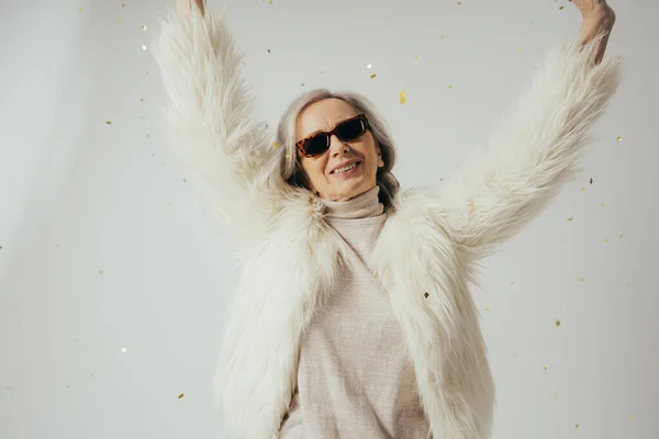 cheerful elderly woman in white faux fur jacket and sunglasses raising hands near falling confetti on grey background