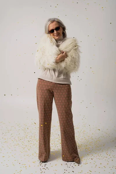 full length of cheerful elderly woman in white faux fur jacket and sunglasses standing near falling confetti on grey background