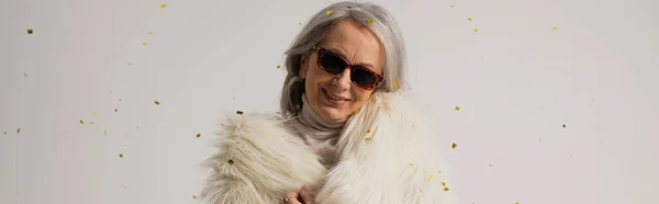 pleased elderly woman in white faux fur jacket and sunglasses smiling near falling confetti on grey background, banner
