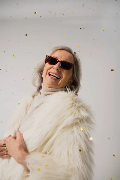 cheerful elderly woman in white faux fur jacket and trendy sunglasses laughing near falling confetti on grey background
