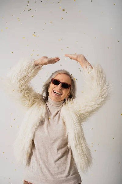 happy elderly woman in white faux fur jacket and sunglasses standing with raised hands near falling confetti on grey