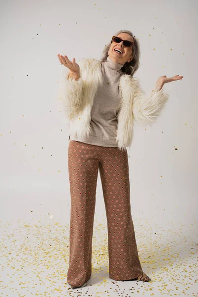 full length of happy elderly woman in white faux fur jacket and trendy sunglasses smiling near falling confetti on grey