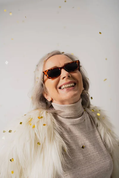 smiling elderly woman in white faux fur jacket and trendy sunglasses near falling confetti on grey background