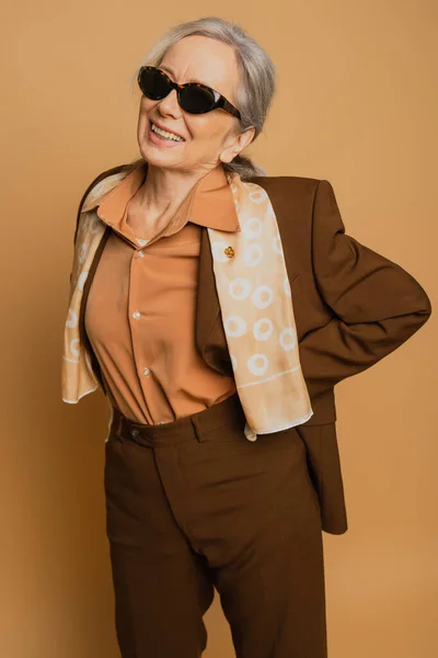 stock image positive and senior woman in sunglasses and suit posing with hand on hip on beige 