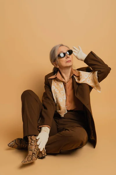 full length of senior woman in suit sitting and adjusting sunglasses on beige background