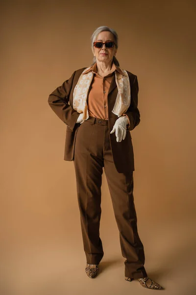 stock image full length of senior woman in brown suit and white gloves posing with hand on hip on beige 