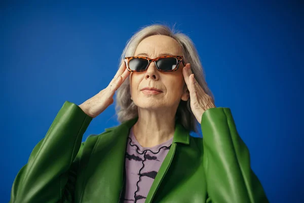 stock image elderly woman in green leather jacket adjusting trendy sunglasses isolated on blue