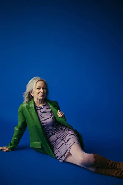 stock image fashionable senior woman in green leather jacket over purple dress smiling at camera while sitting on blue background