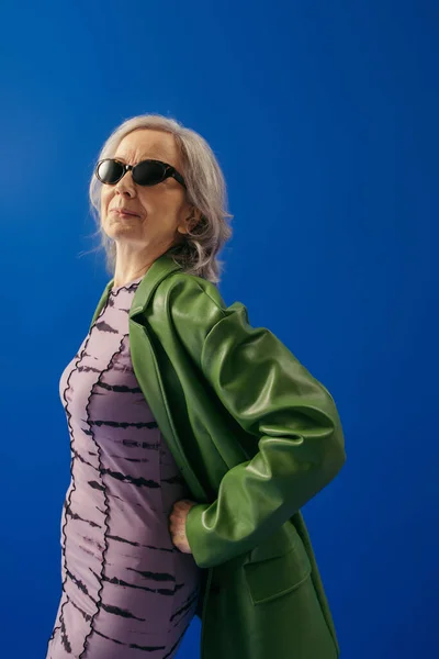 grey haired woman in trendy sunglasses and green leather jacket over purple dress posing with hand on hip isolated on blue