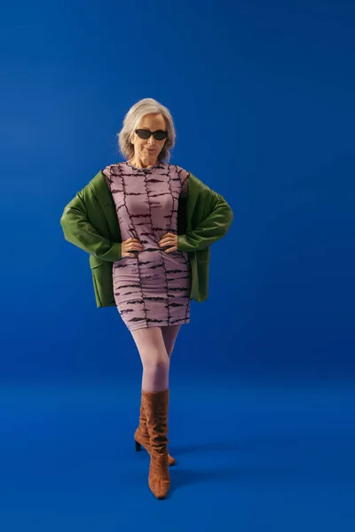 full length of senior lady in elegant dress and green leather jacket posing with hands on waist on blue background