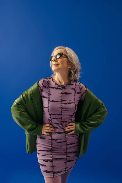 senior model in sunglasses and fashionable outfit posing with hands on waist isolated on blue
