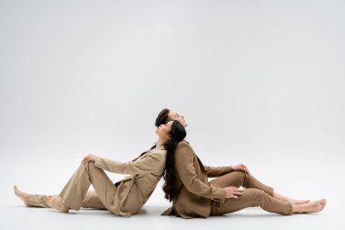 side view of multiethnic couple in beige suits sitting back to back on grey background