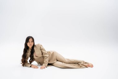full length of barefoot asian model in fashionable suit lying and looking at camera on grey background