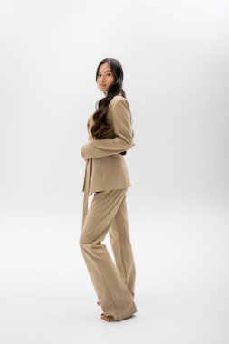 full length of barefoot asian woman in beige jacket and trousers standing on grey background
