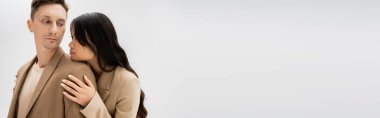 brunette asian woman hugging trendy man in beige blazer isolated on grey, banner
