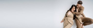 high angle view of man embracing sensual asian woman while sitting in trendy outfit on grey background, banner