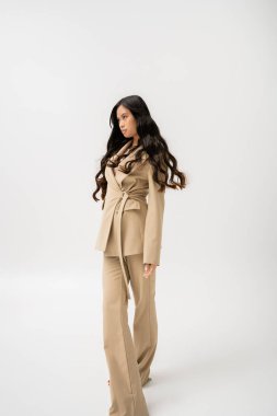 long haired asian woman in beige pantsuit looking away while standing on grey background