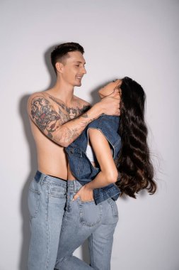 smiling man with shirtless tattooed body embracing asian woman with long brunette hair on grey background