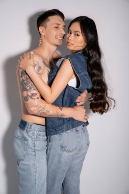 pleased man with shirtless tattooed body hugging brunette asian woman in denim outfit on grey background