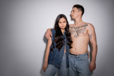 brunette asian woman in denim vest and jeans looking at camera near shirtless tattooed man on grey background