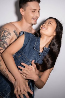shirtless tattooed man embracing joyful asian woman in denim vest on grey background