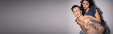 tattooed and muscular man smiling at camera while piggybacking asian woman on grey background, banner