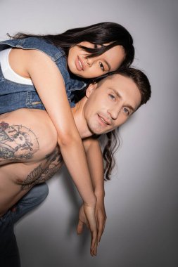 shirtless tattooed man and asian woman in denim vest looking at camera while having fun on grey background