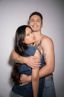cheerful tattooed man embracing stylish asian woman pouting lips on grey background
