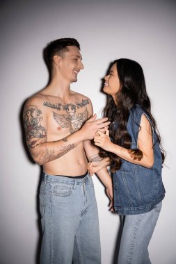 cheerful asian woman and shirtless tattooed man smiling at each other on grey background